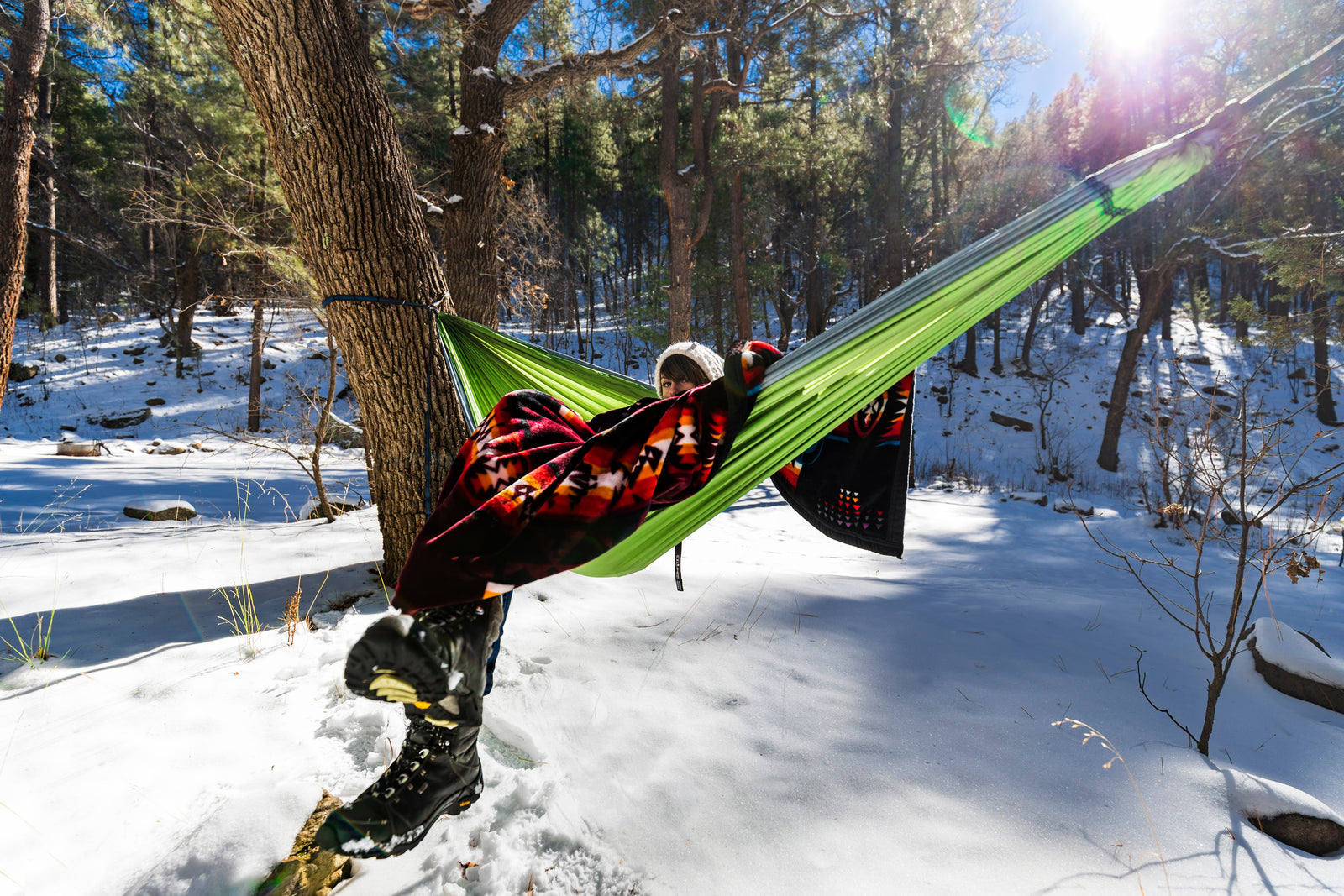 A woman bundled up in a red blanket sitting in a hammock in a snowy forest.