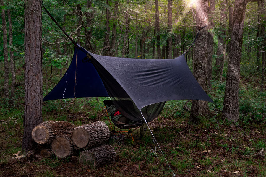 Hammock covered by hammock tarp hung up in a damp forrest at dawn with logs next to it.