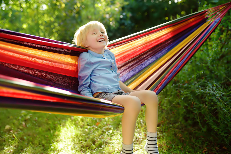 Child sitting in a rainbow hammock on a sunny day, looking happy.