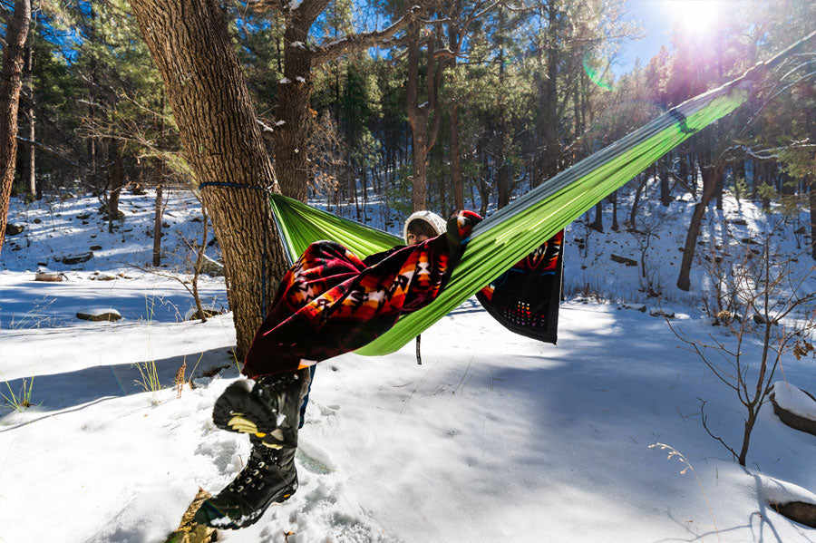 A woman wrapped in a red blanket sitting on a hammock in a snowy, sunlit forest.