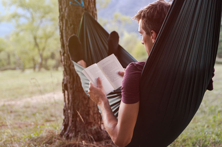 A man reading a book while laying in a hammock in a park.