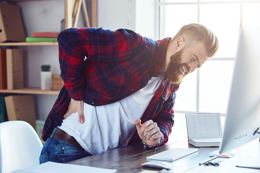 A bearded man hunched over his desk clutching his lower back in pain.