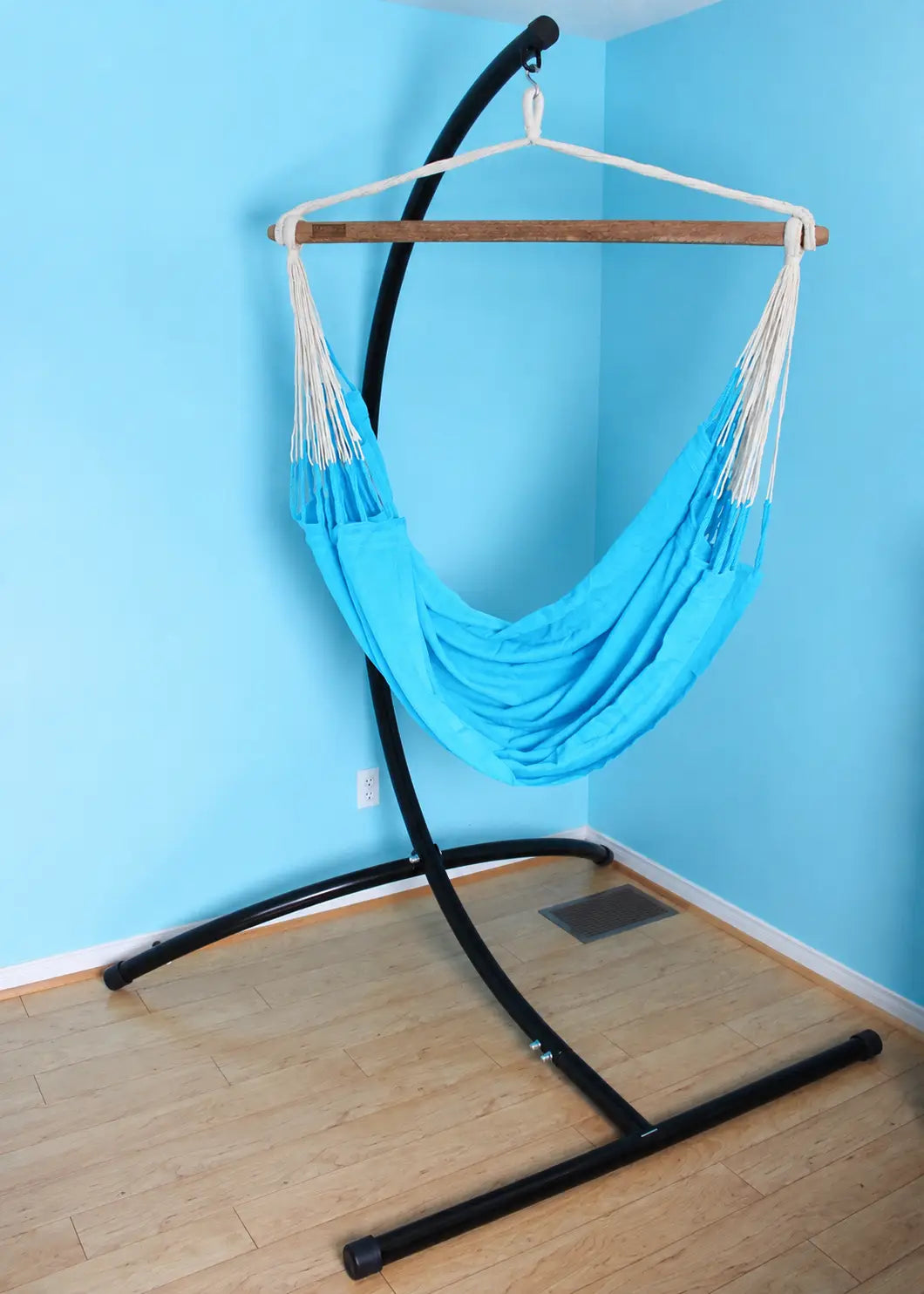 A bright blue fabric hammock suspended from a black metal stand with a wooden hanging bar, set against a light blue wall and light wood floor.