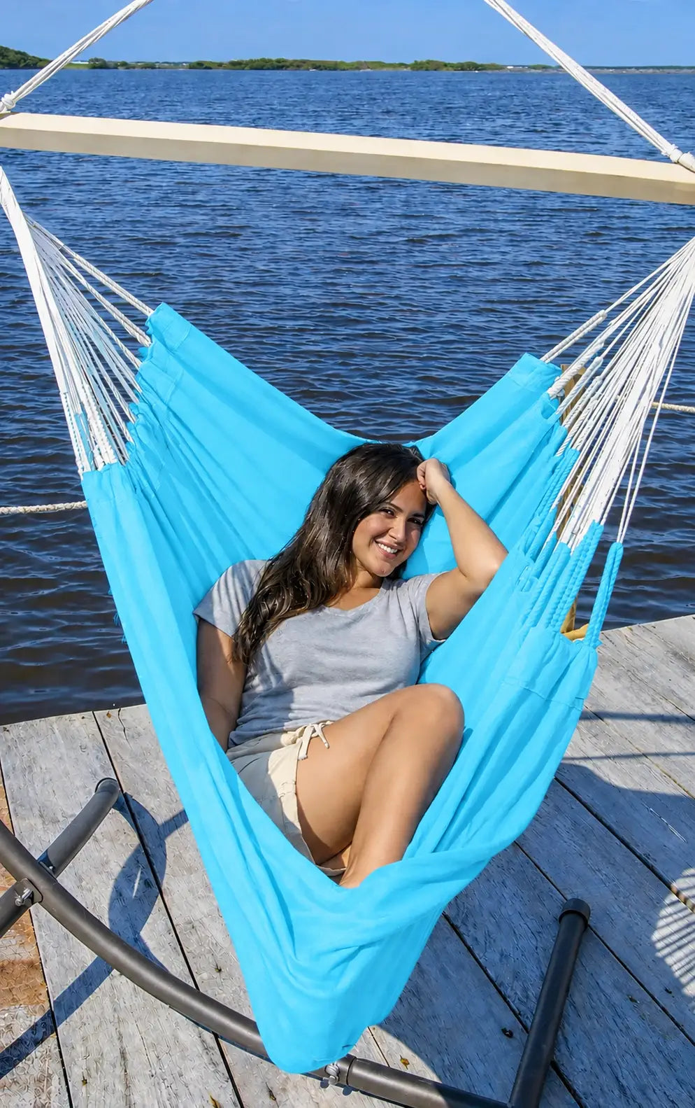 A woman relaxes in a bright turquoise blue hammock suspended over a wooden dock with blue water in the background.