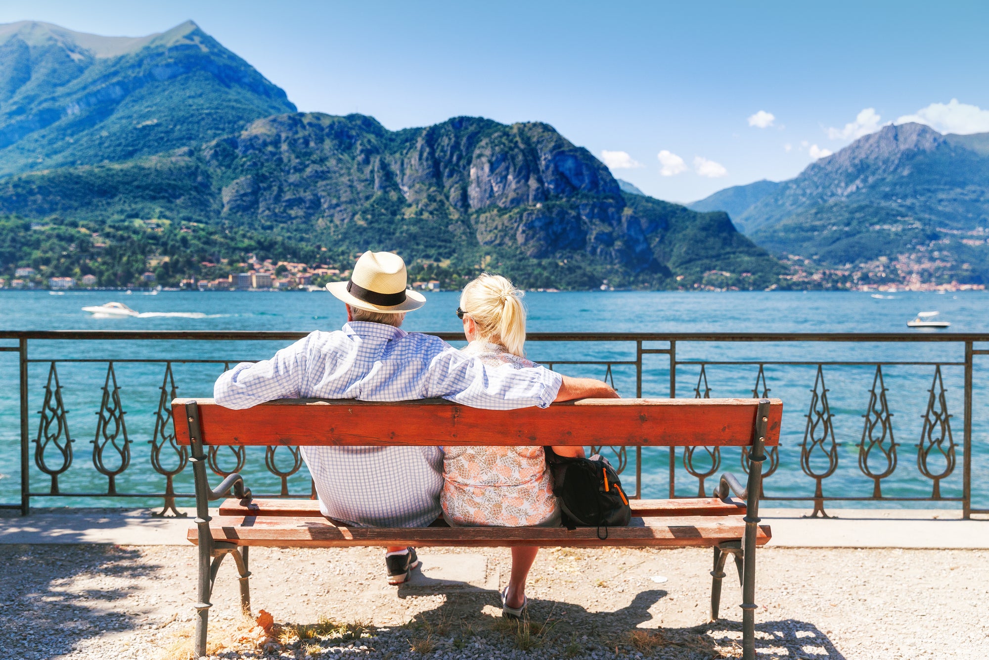 A couple sitting at a bench facing the mountains and a lake.