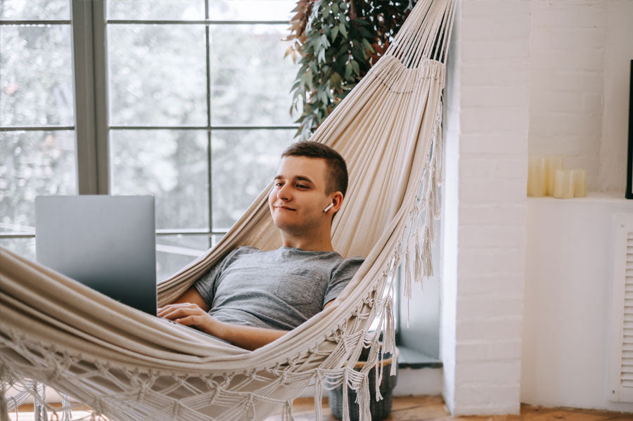 A man watching something on his laptop while lounging in his indoor hammock.
