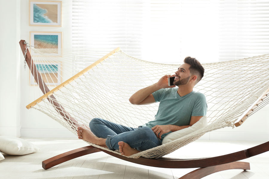 A smiling man on the phone, sitting on a rope hammock with stand in a modern room.