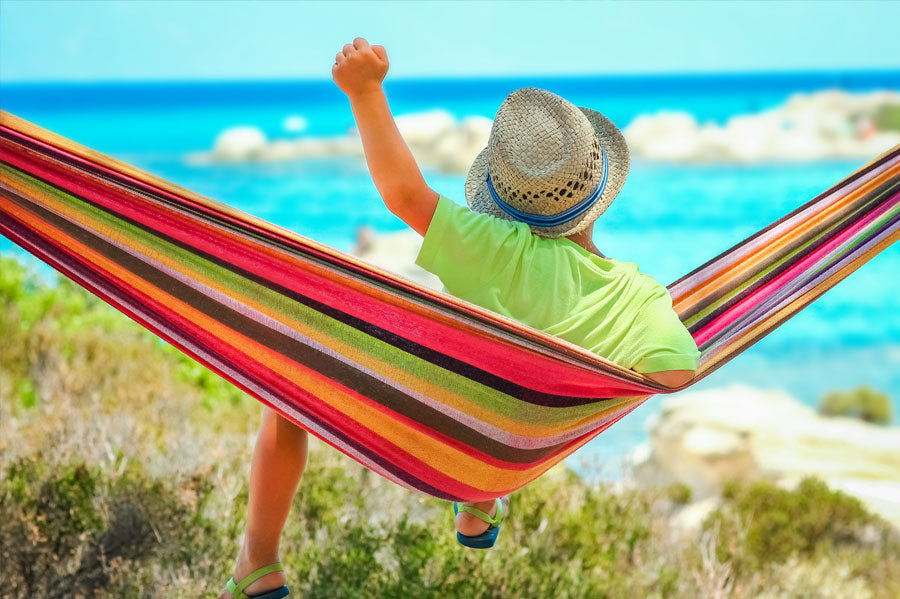 A child lounging and raising his hand in a hammock at a beach.