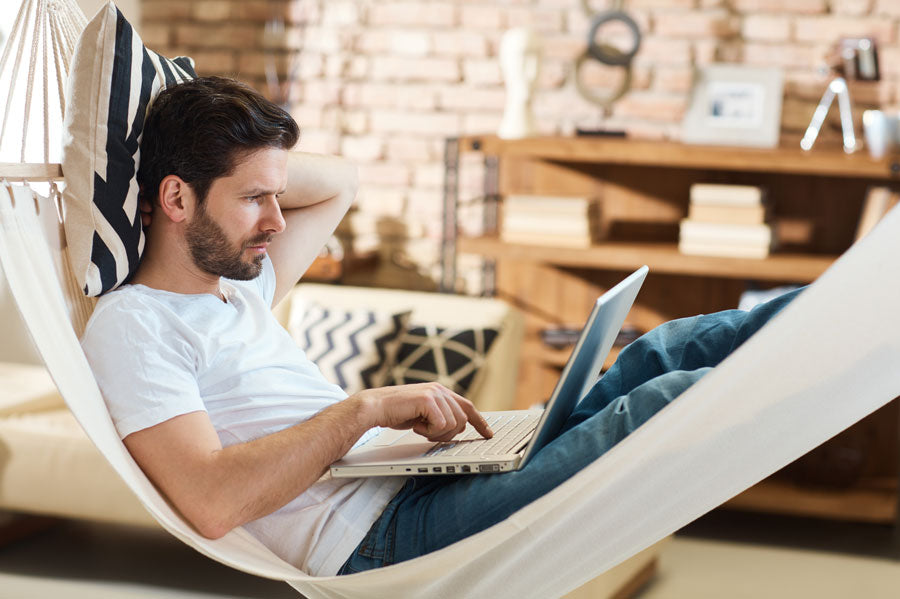 A man working on his laptop on a hammock in his home.