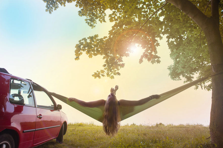 A woman laying in a hammock that is hung up from a tree and her car at sunset.