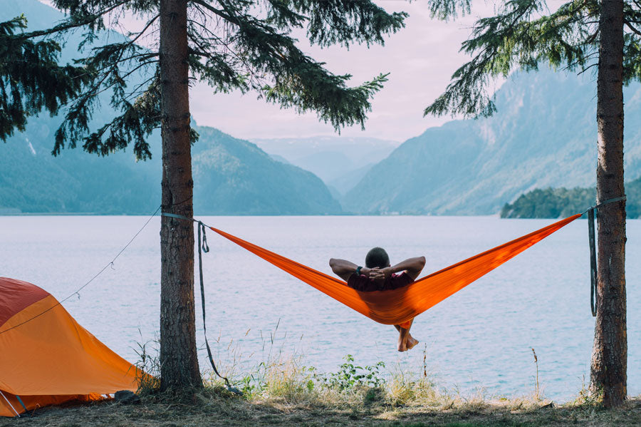 A man lounging in a hammock hung in between two trees in a lake side campsite.