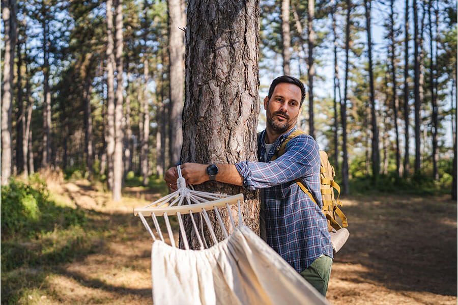 A hiker secures a hammock to a sturdy tree in a wooded area, preparing for a relaxing outdoor experience.