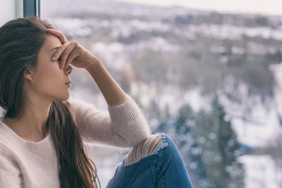 An unhappy woman with her hand over her eyes, sitting at a window.