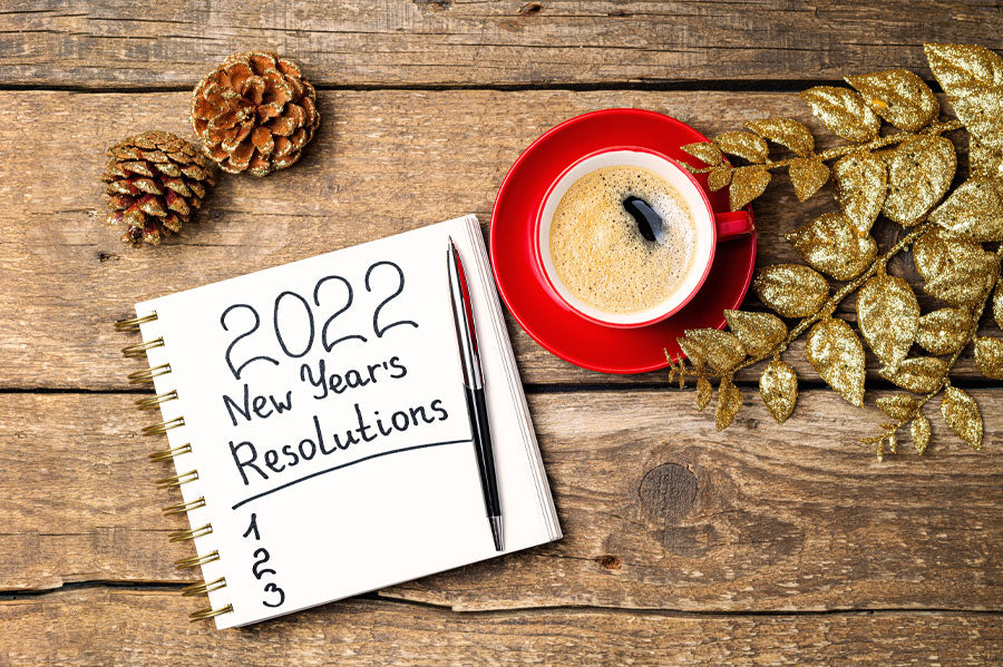 Top view of a notebook with "2022 New Years Resolutions" written on it next to a coffee cup with gold leaves and pine cones around them.