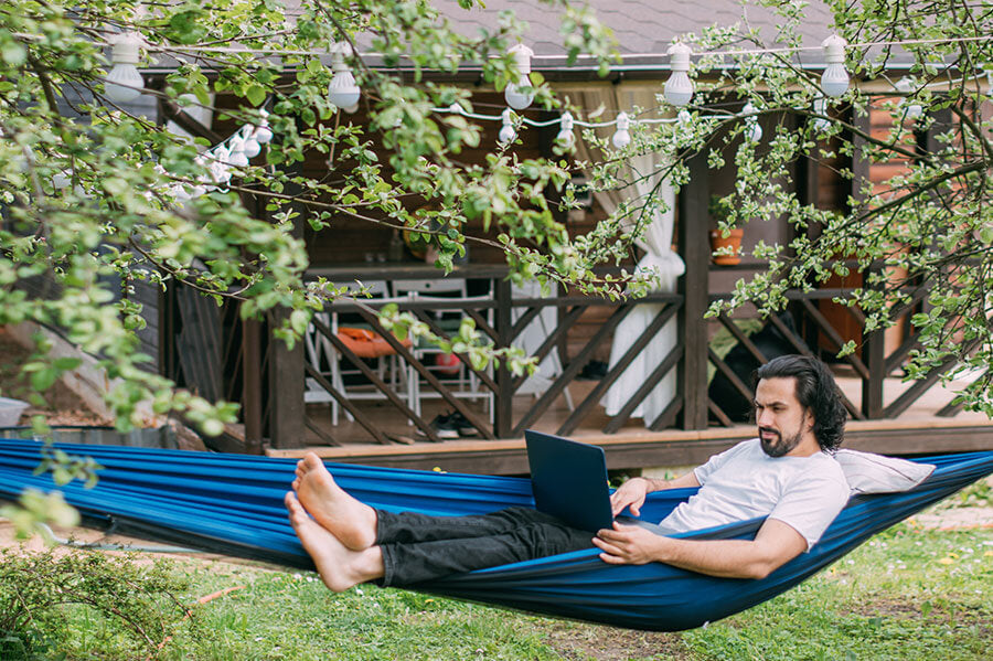 A man relaxing in his hammock in the backyard, working on his laptop while enjoying the outdoors.