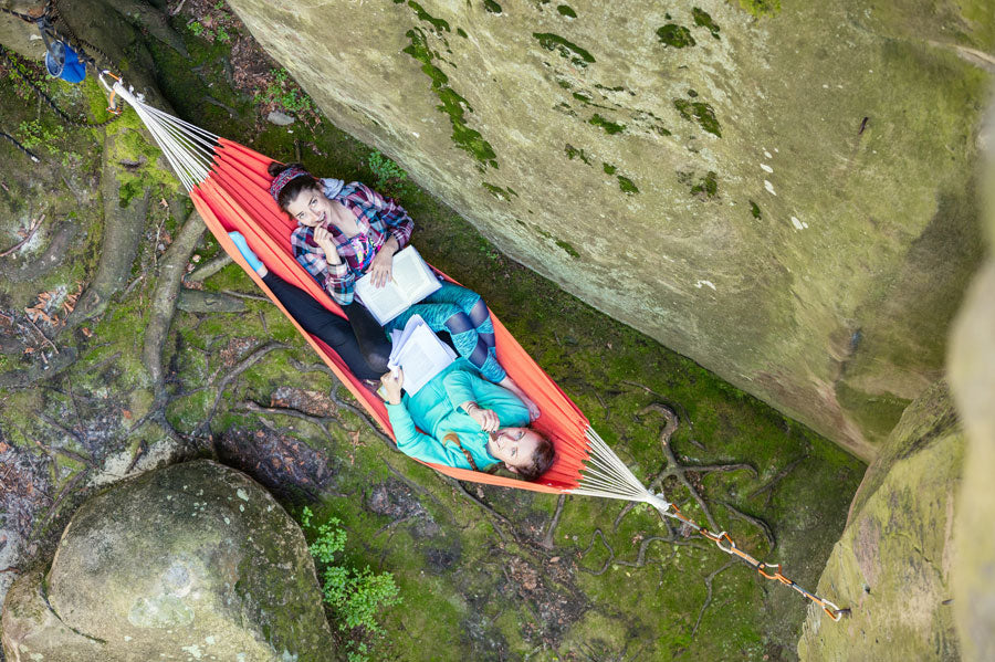 Top view of two women eating a snack and reading while laying in a hammock hung on a cliff side.