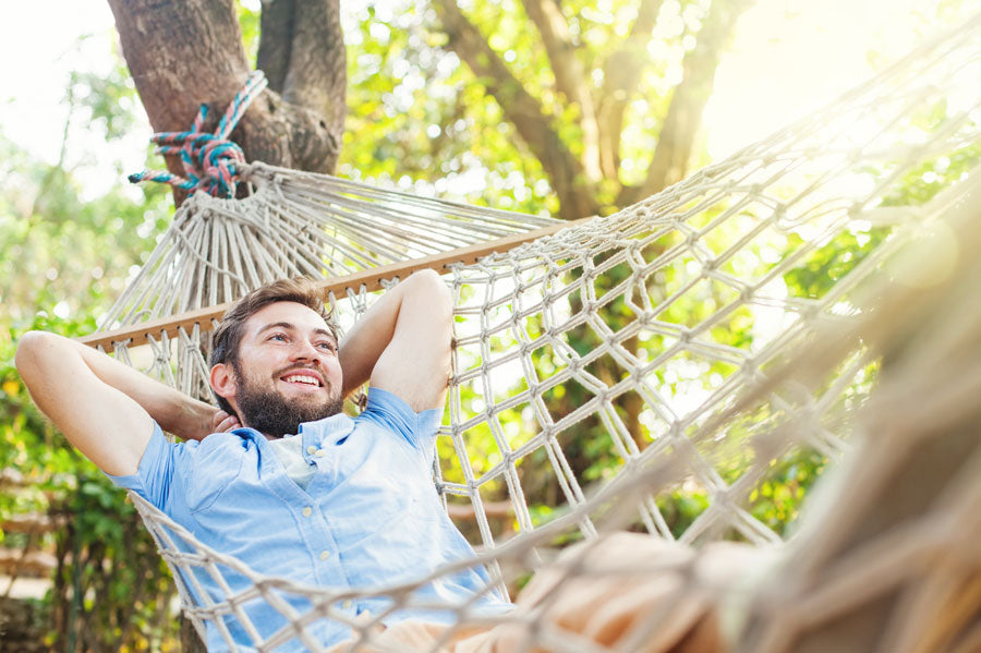 A smiling man relaxing in a rope hammock in sunny woods.