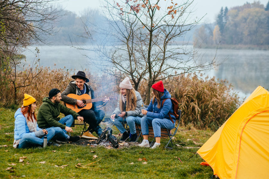 A group of millennials camping and sitting around a camp fire smiling and listening to the guitar.