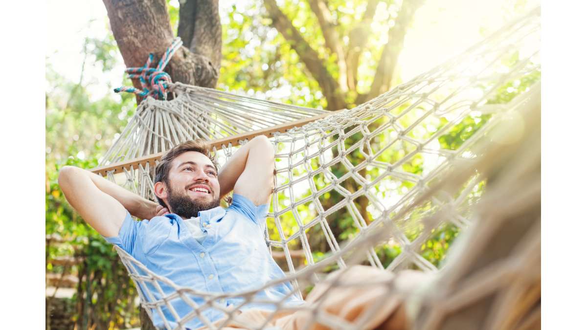 A smiling man lounging in a rope hammock in sunlit woods.
