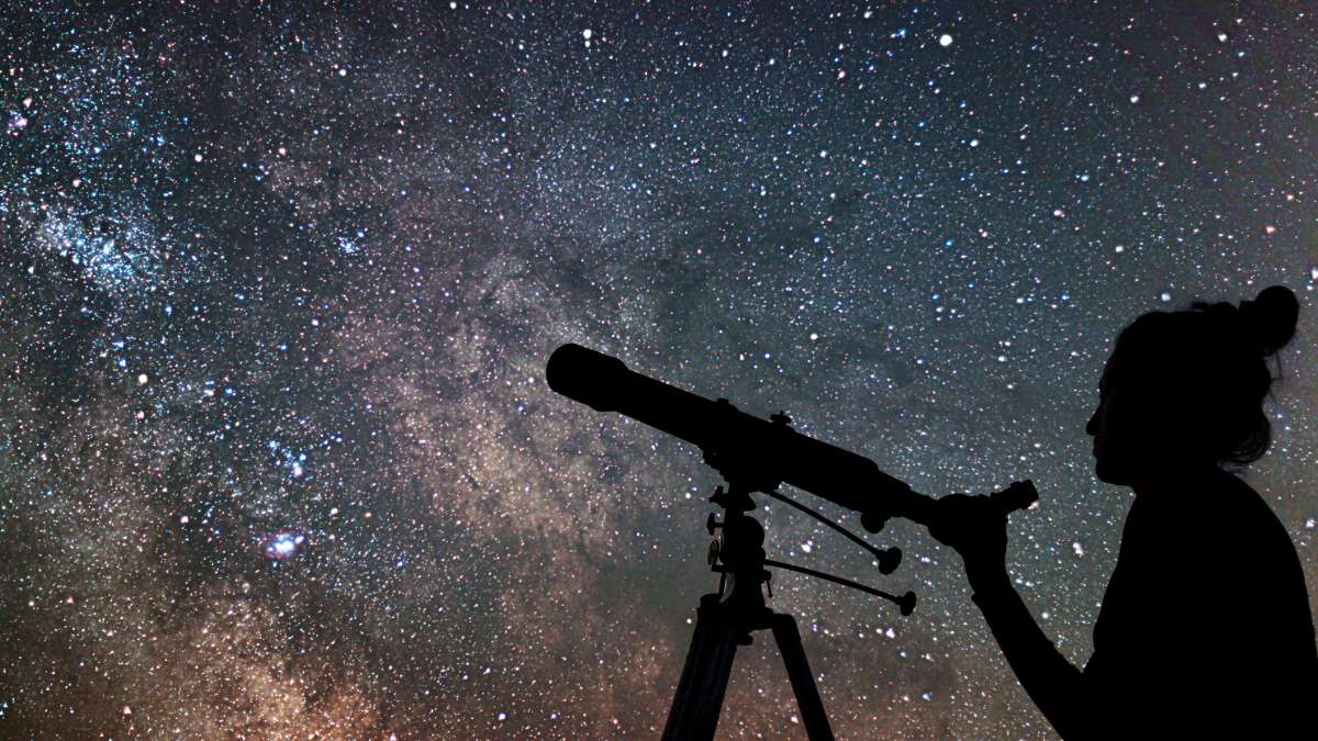 A silhouette of a woman looking in a telescope in front of a starry night sky.
