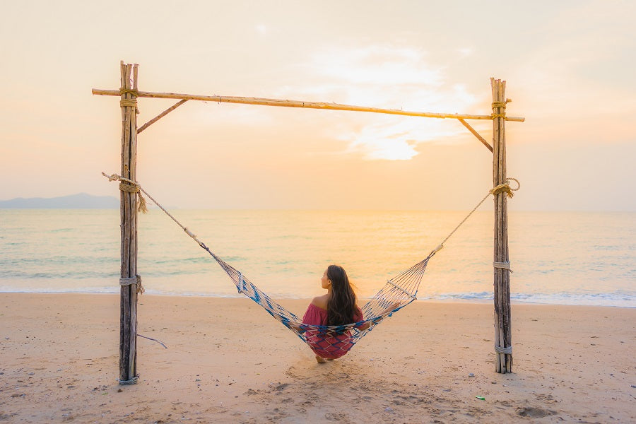A woman sitting in a hammock on the beach, facing the ocean and sunset.