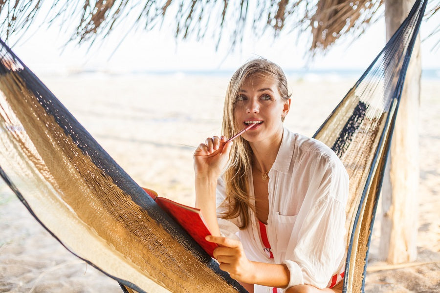 A woman holding a pen in her mouth while sitting in a hammock on the beach.