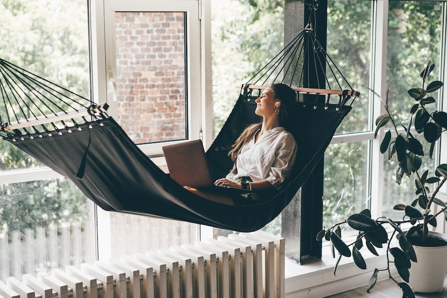 a woman lying in her hammock with a laptop in her lap, indoor setting next to the window