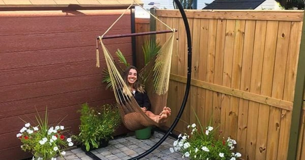 A young woman sits in a Mayan hammock chair with universal stand in her backyard next to plants and flowers.