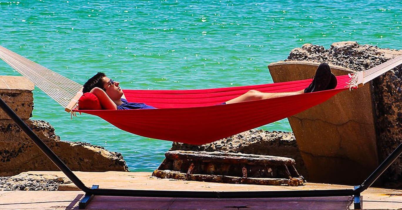 A man lounging in a red hammock with stand on a sunny day in front of the ocean.