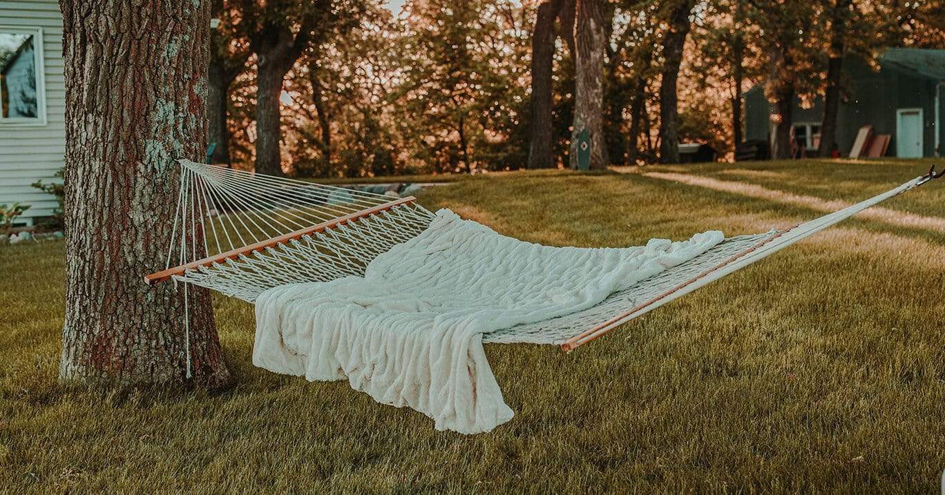 A cotton rope hammock with spreader bars hangs between trees in the backyard with a cozy blanket on it