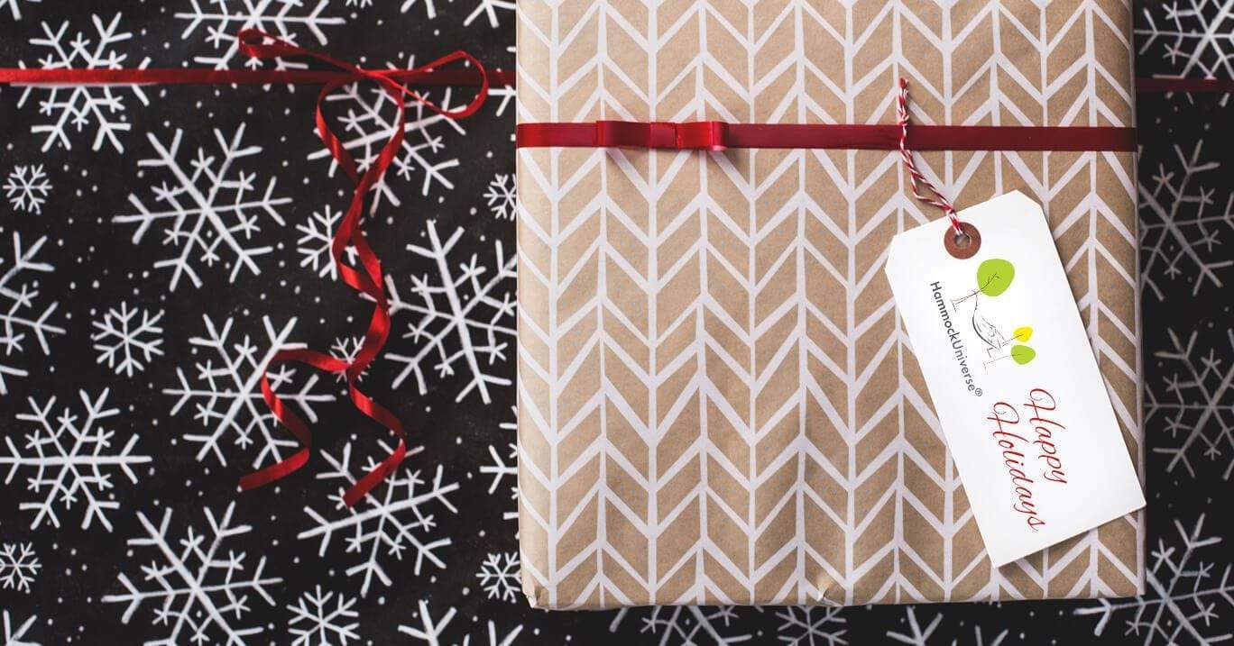 Closeup of two wrapped gifts, the on top has a tag saying "Happy Holidays" next to the Hammock Universe logo.