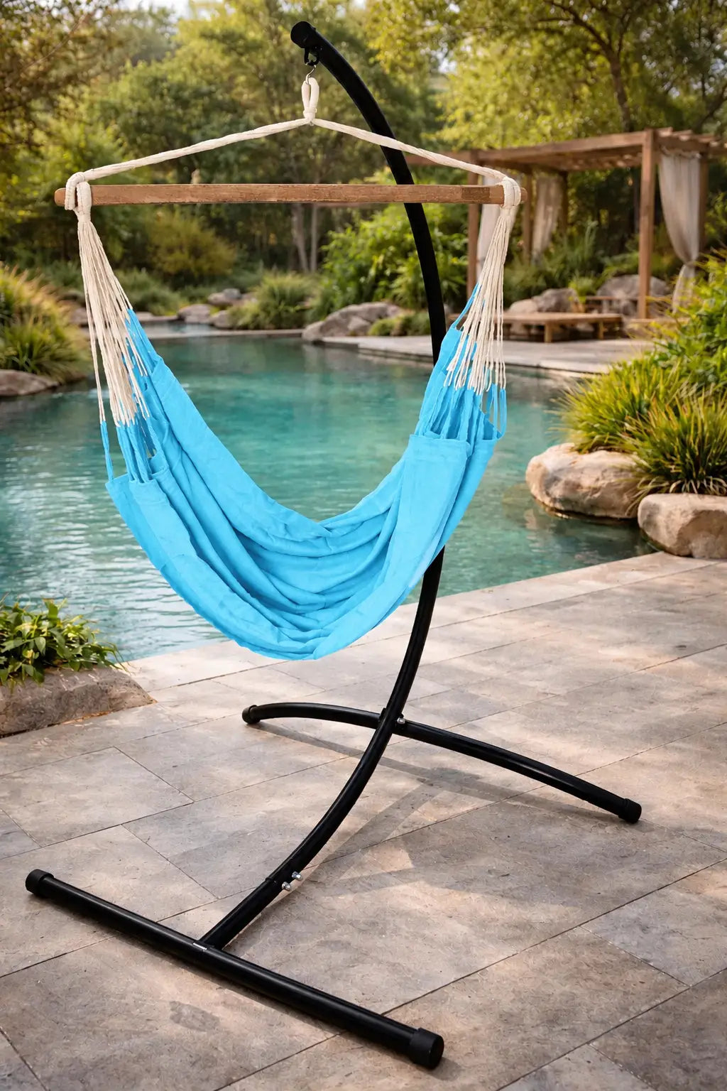 A bright blue fabric hammock suspended on a black metal stand, set on a stone patio beside a swimming pool with green landscaping in the background.