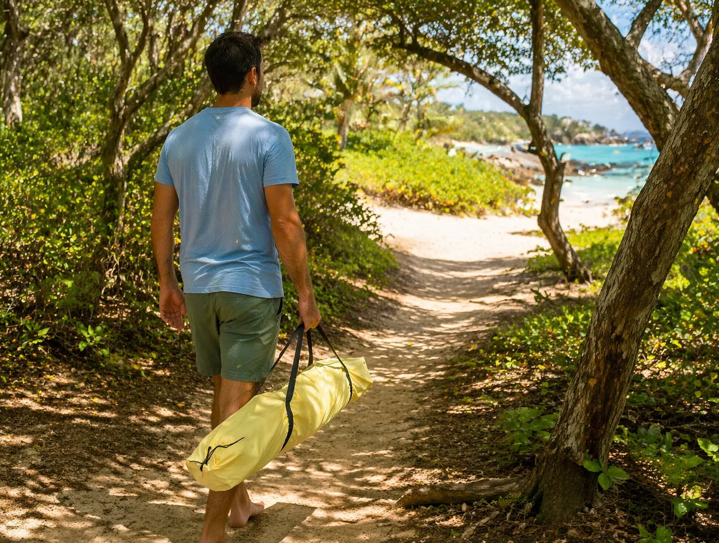 Man walking along a trail with a yellow bag, surrounded by greenery and a beach in the distance.