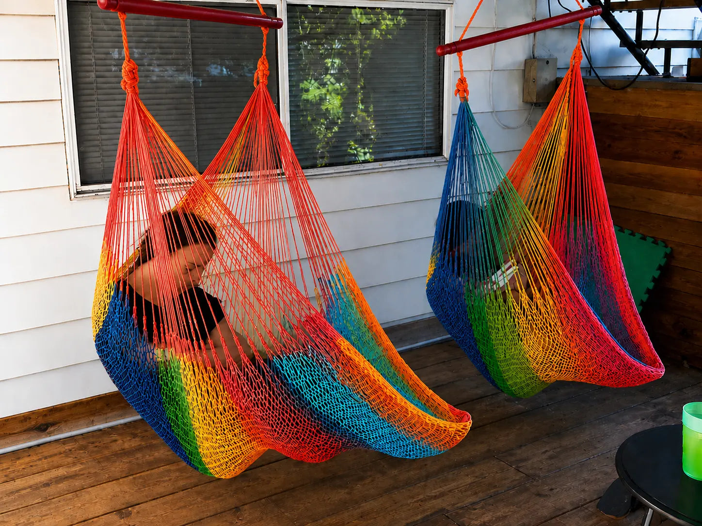 Child sitting in a colorful hammock on a wooden floor with a window in the background.