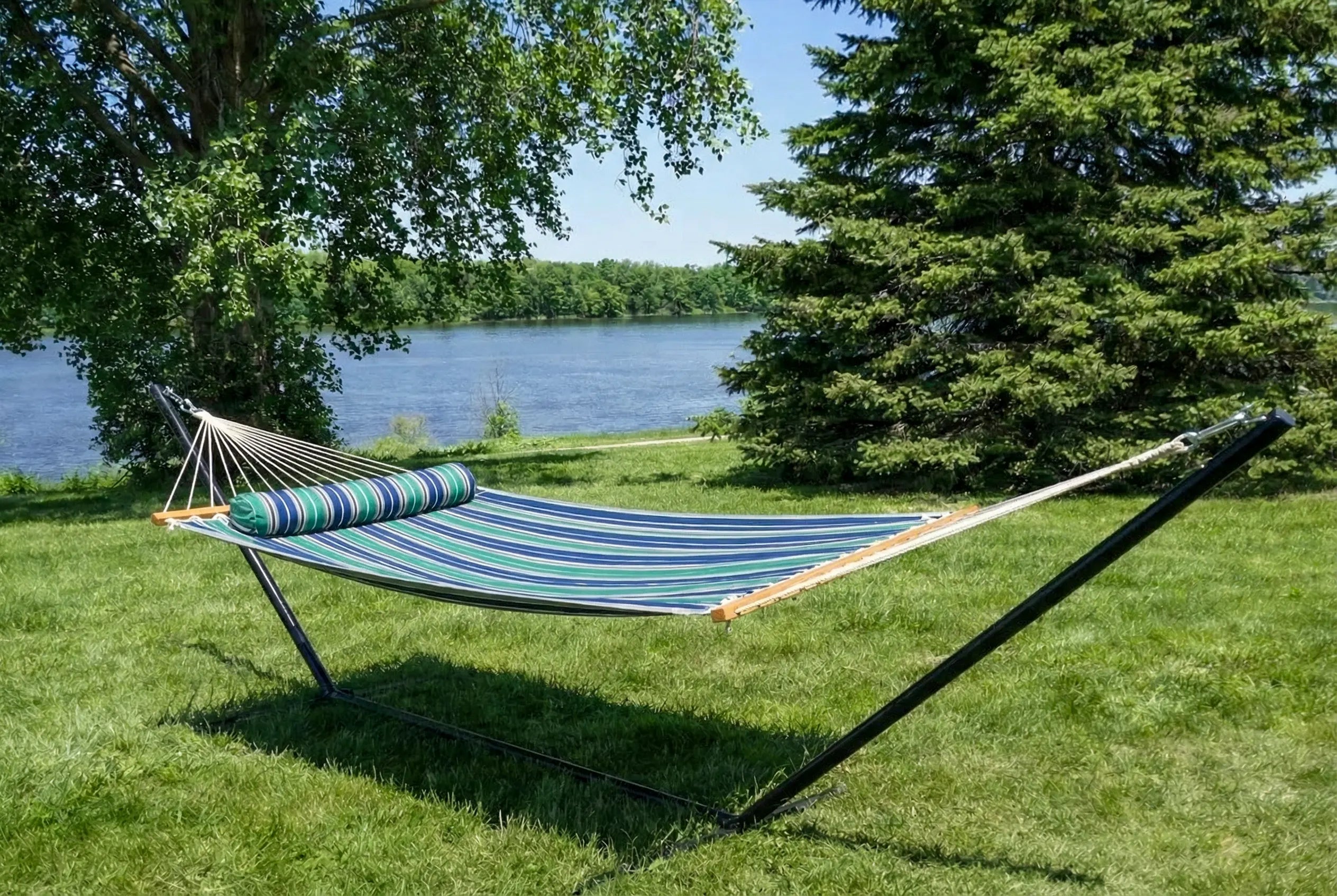 Striped hammock on a stand in a grassy area near a body of water with trees.  QHD-GBGW+15TBSB