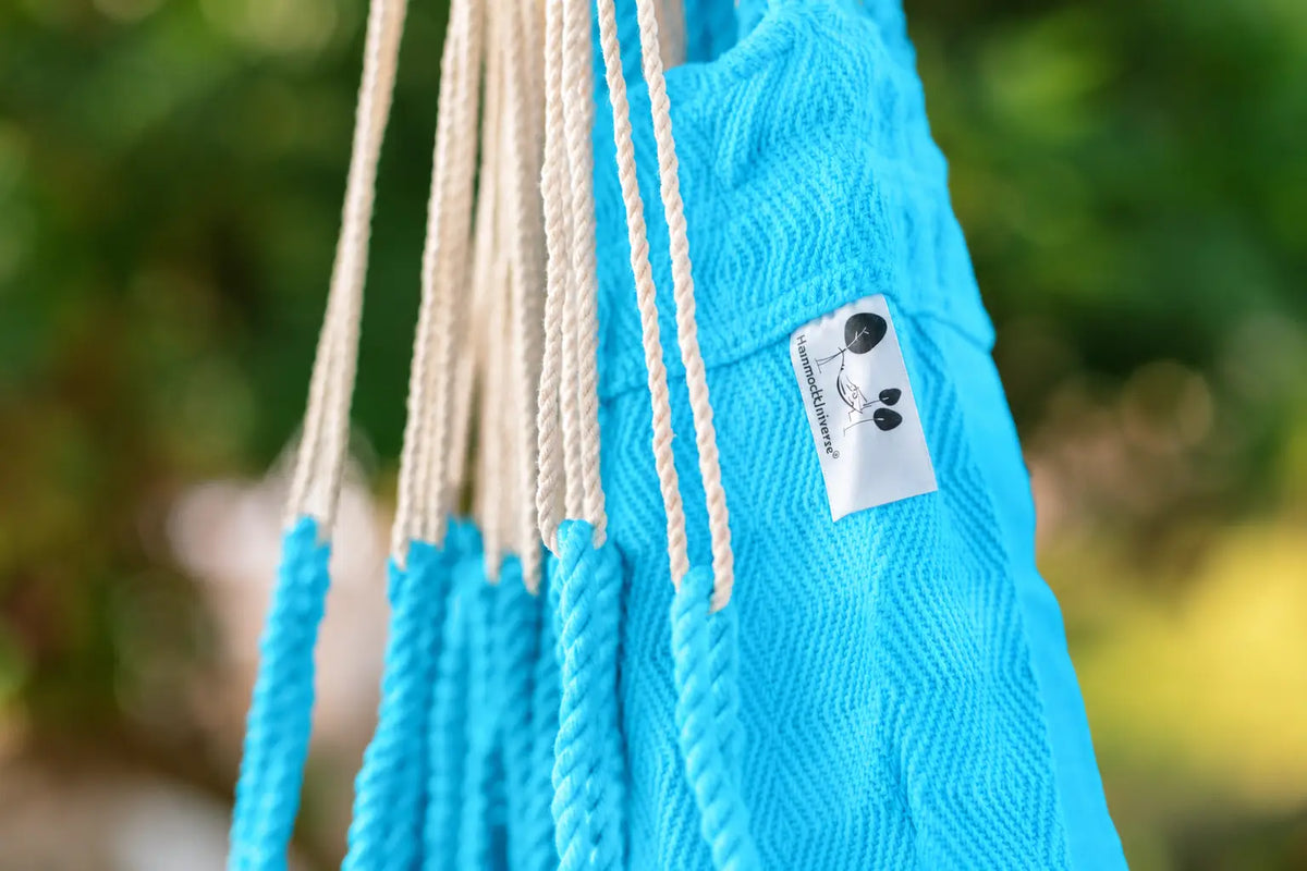 A close-up of a bright blue woven hammock with cream-colored hanging ropes and a small white brand tag that reads 'Panda's Paradise'. The background is a soft, blurred green outdoor setting.