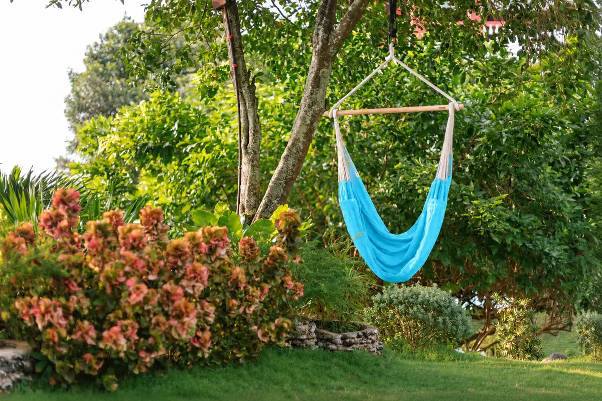 A bright blue hammock with beige fabric sides and a wooden spreader bar hangs from a tree in a lush green garden with flowers and grass.